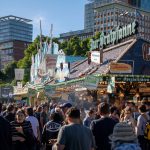 Público circula por um festival gastronômico ao ar livre, entre barracas de comida, sob um céu azul.