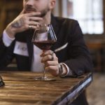 Homem de terno e crachá segura uma taça de vinho diante de uma mesa de degustação em ambiente interno.