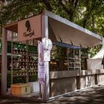 Barraca de vinhos em um mercado ao ar livre, cercada por árvores e movimentação de visitantes.