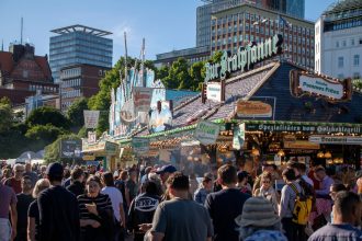 Público circula por um festival gastronômico ao ar livre, entre barracas de comida, sob um céu azul.