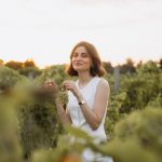 Mulher sorridente segura um cacho de uvas em um vinhedo ao pôr do sol.