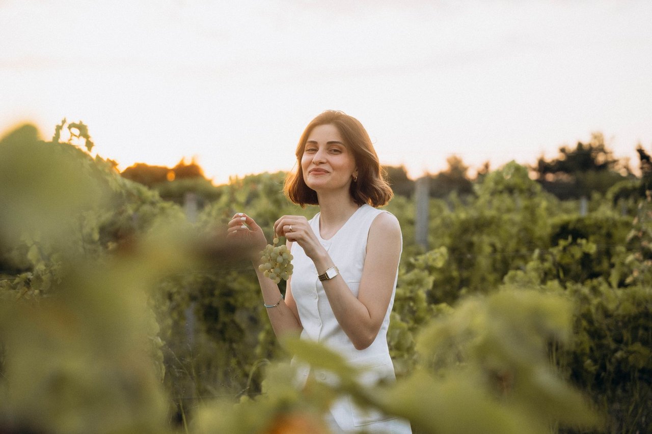Mulher sorridente segura um cacho de uvas em um vinhedo ao pôr do sol.